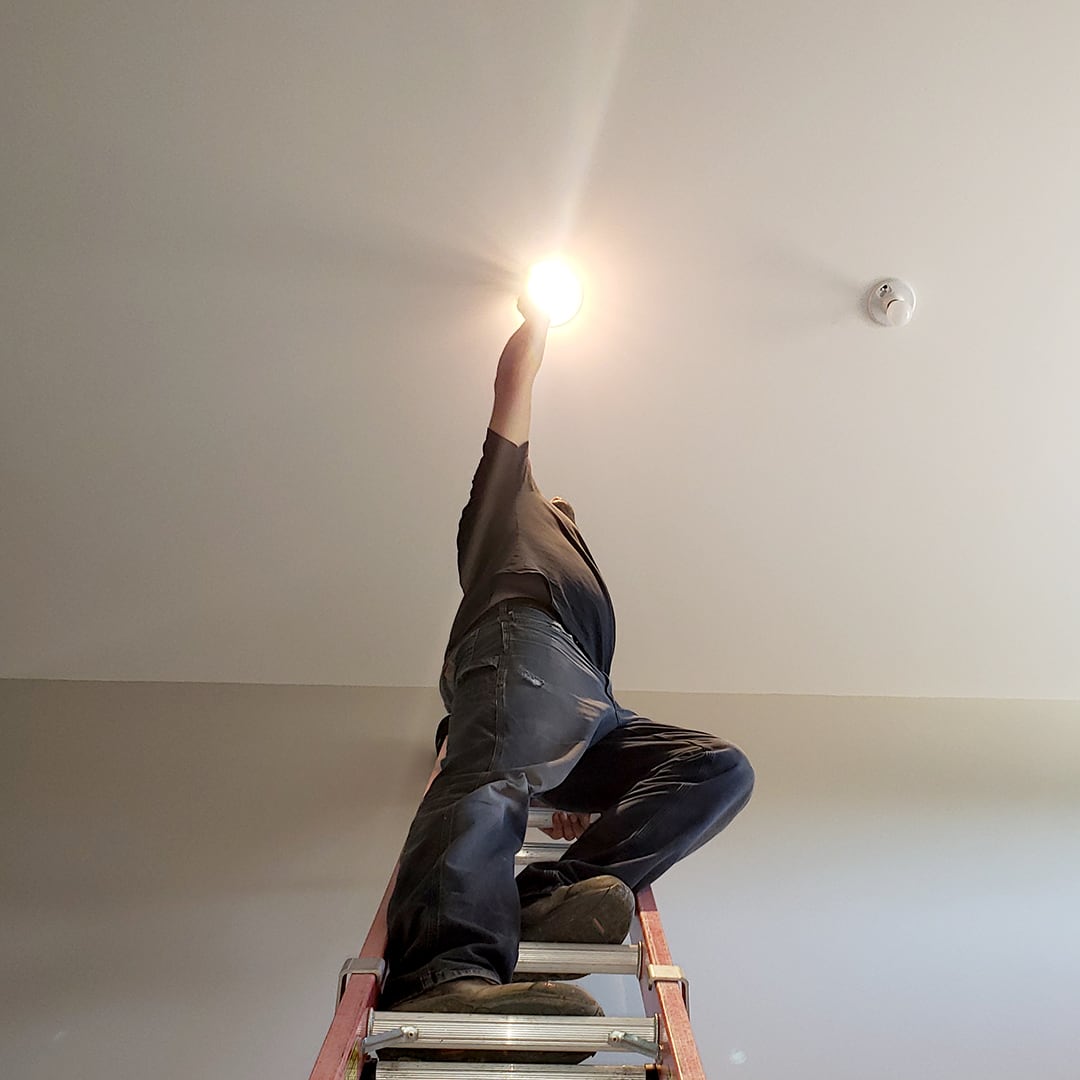 Low-angle view of an electrician on a ladder, looking up and changing a lightbulb, with a bright lens flare near his outstretched hand
