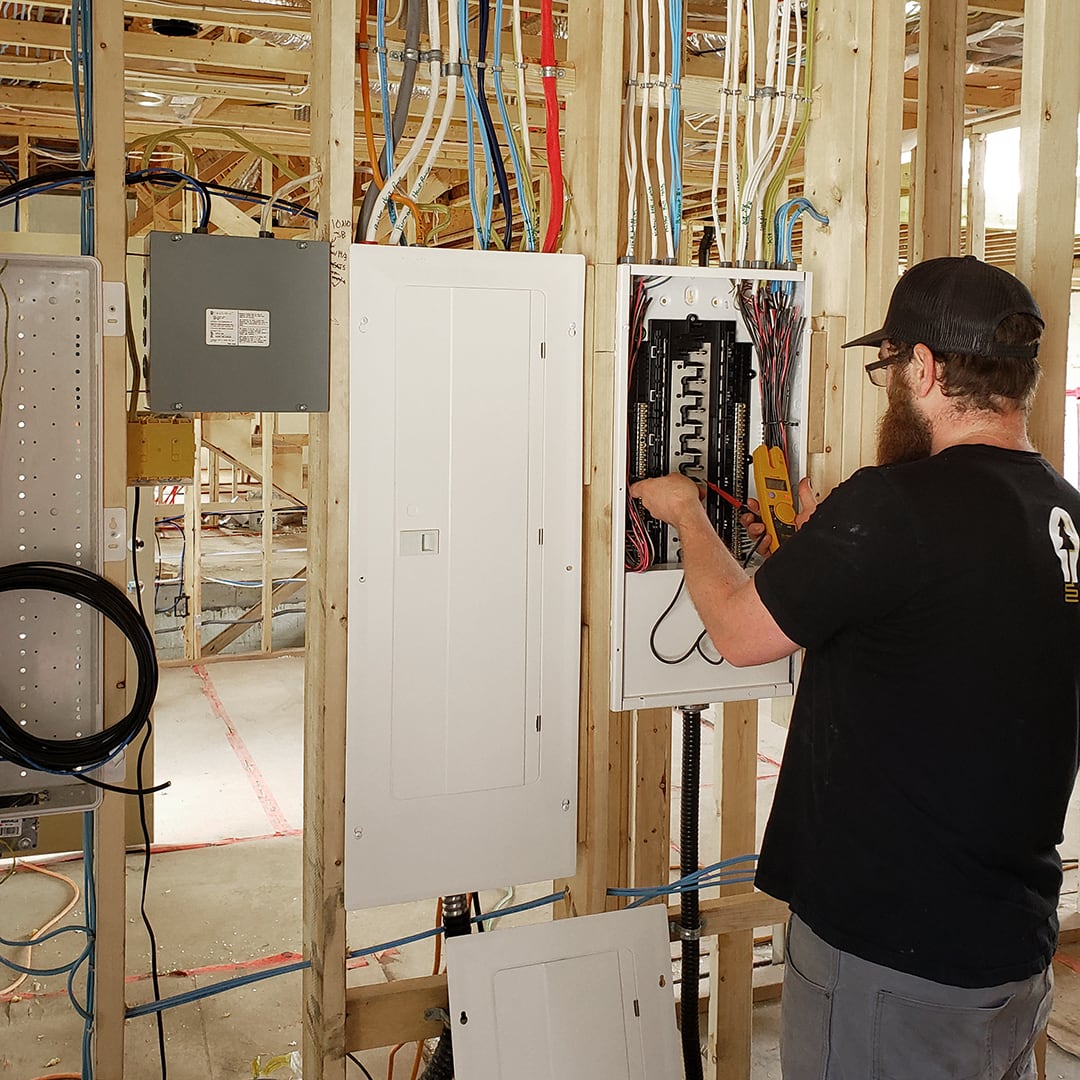 Electrician with his back to the camera, wearing a black shirt and hat with the Island Electric Co. logo on it, working on a residential service panel in a new construction project