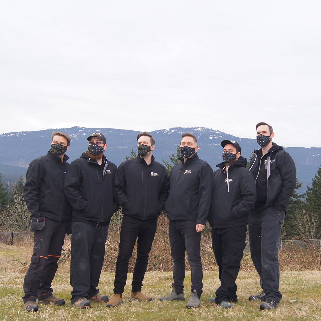 Group shot of the Island Electric Company electricians, standing together in a field in front of Mt Benson in Nanaimo, BC, wearing black face coverings