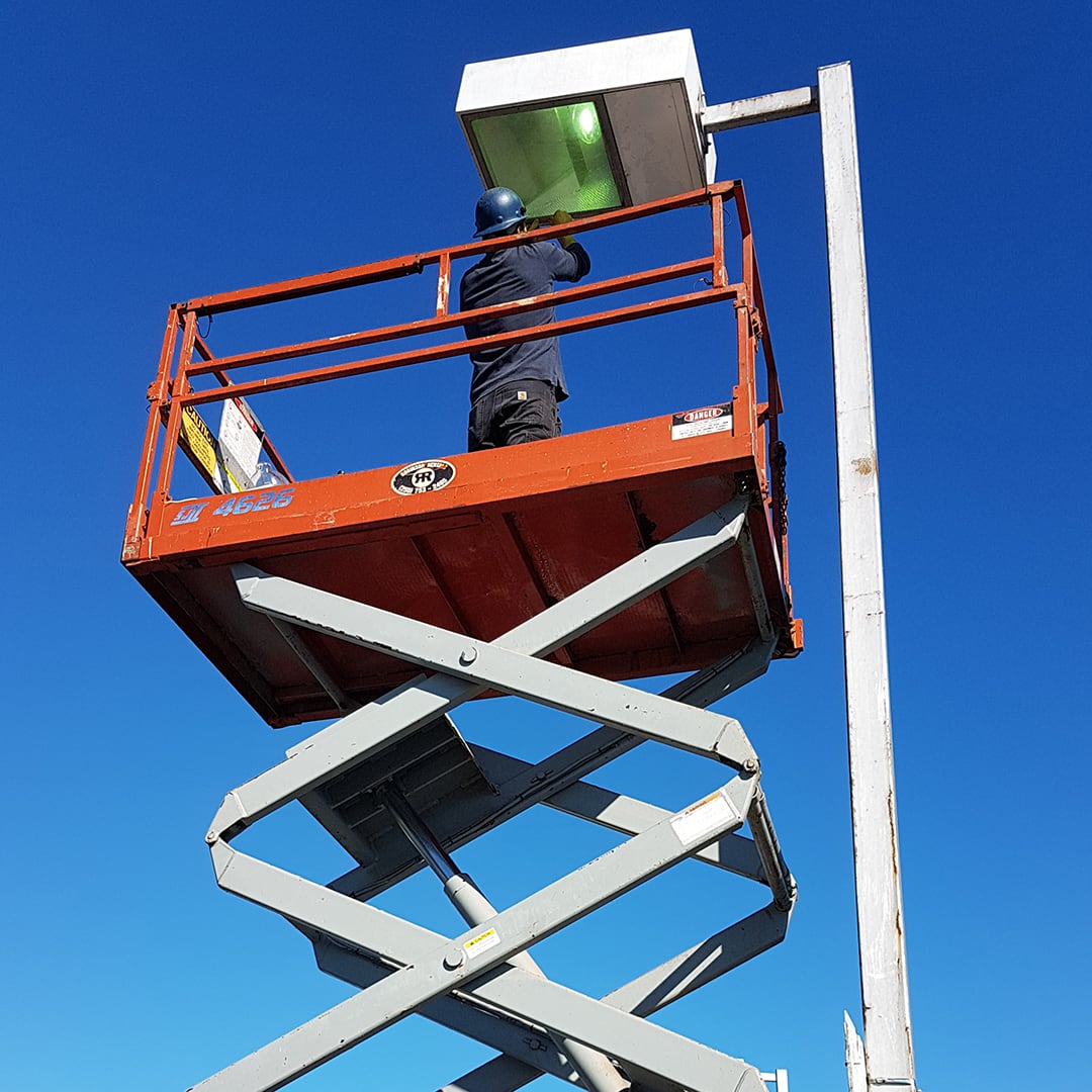 Red scissor-lift platform with an electrician on top, arms stretched above him as he works on a parking lot lamp