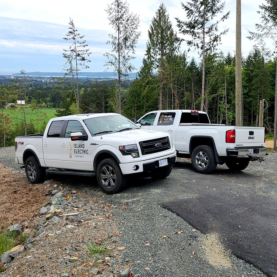 Two white pickup trucks with the Island Electric Company logos on the door and rear windows, parked at an angle on a residential road that is under construction